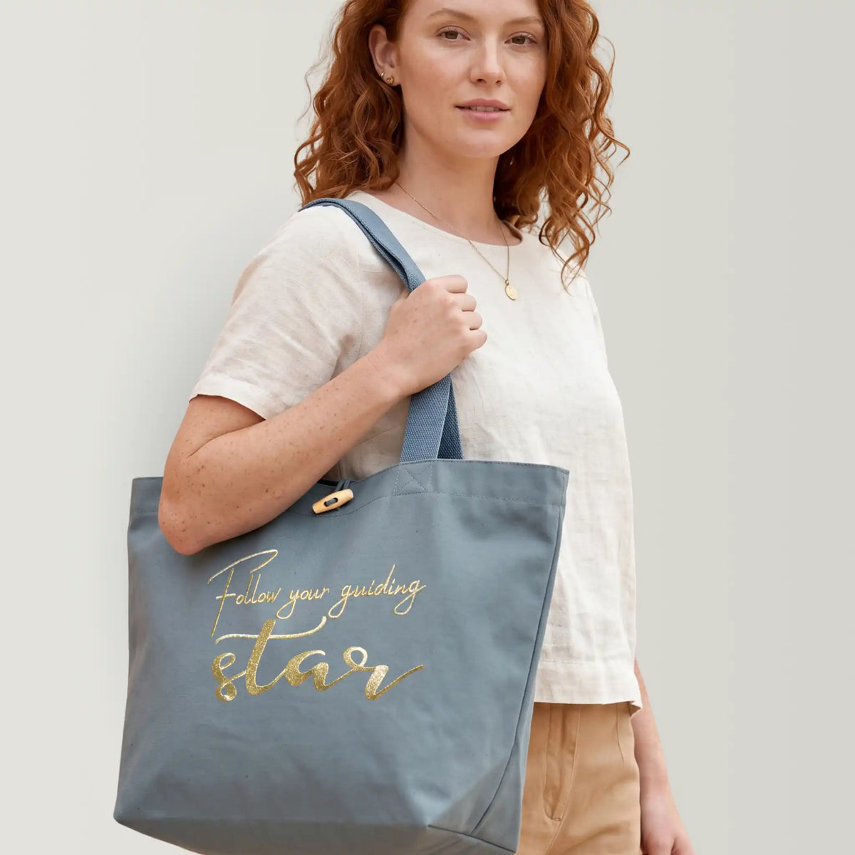 Model trägt große hellblaue Tote Bag mit Affirmation als schimmernde Goldstickerei. Stickereitext: "Follow your guiding star". Knebelverschluss, Tote Bag aus robustem Canvas und 100% Biobaumwolle