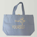 Flatlay einer robusten hellblauen XL GOTS-Tote Bag aus stabilem Canvas (100% Biobaumwolle) mit langen Henkeln, große Stickerei in gold-metallic Schriftzug Trust yourself mit minimalistischen floralen Design.