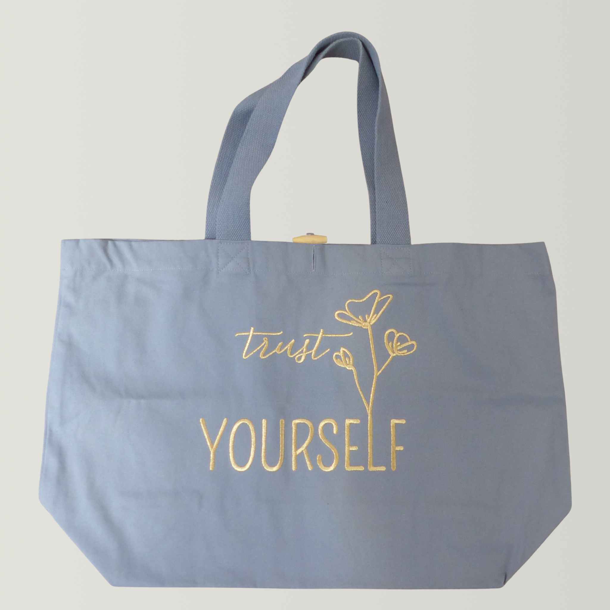 Flatlay einer robusten hellblauen XL GOTS-Tote Bag aus stabilem Canvas (100% Biobaumwolle) mit langen Henkeln, große Stickerei in gold-metallic Schriftzug Trust yourself mit minimalistischen floralen Design.