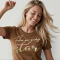 Blondes Model tanzt vor grauer Wand, sie trägt hellbraunes Bio TShirt für Damen der Marke Leitstern, mit golden schimmernder Frontstickerei Schriftzug 'Follow your guiding star'. Logostick am rechten Ärmel.