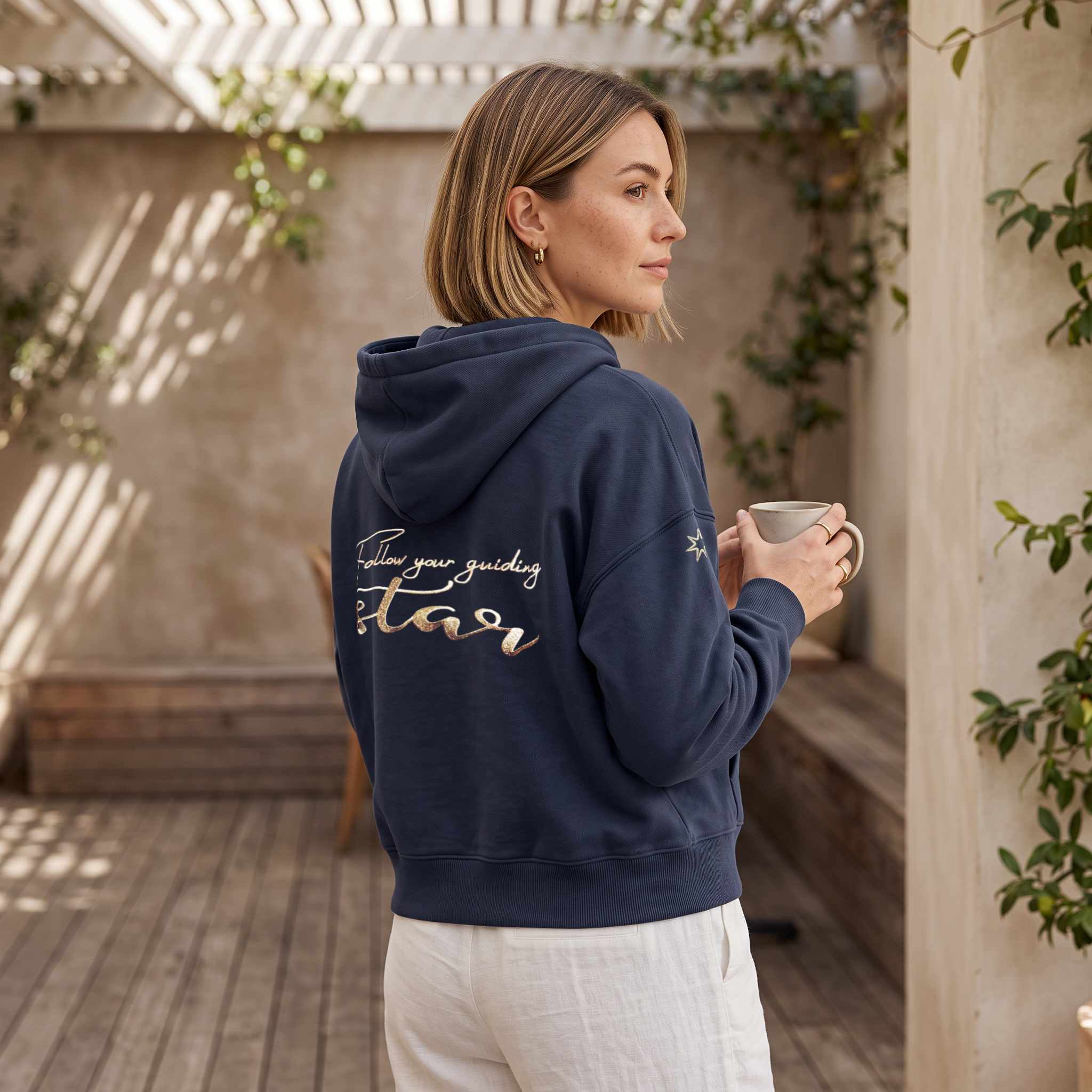 Model trägt eine dunkelblauen Damensweatjacke aus 100% Biobaumwolle mit Kapuze, große Stickerei am Rücken in gold-metallic, Schriftzug Follow your guiding star, Logostick am Ärmel: siebenzackiger Stern