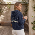 Model trägt eine dunkelblaue Damen Baumwoll Sweatjacke mit Affirmation, große Stickerei am Rücken in metallic-Gold, Affirmation 'Follow your guiding star'. Logostickerei am Ärmel in metallic-gold: siebenzackiger Stern. Sie steht auf einer schönen Terrasse.