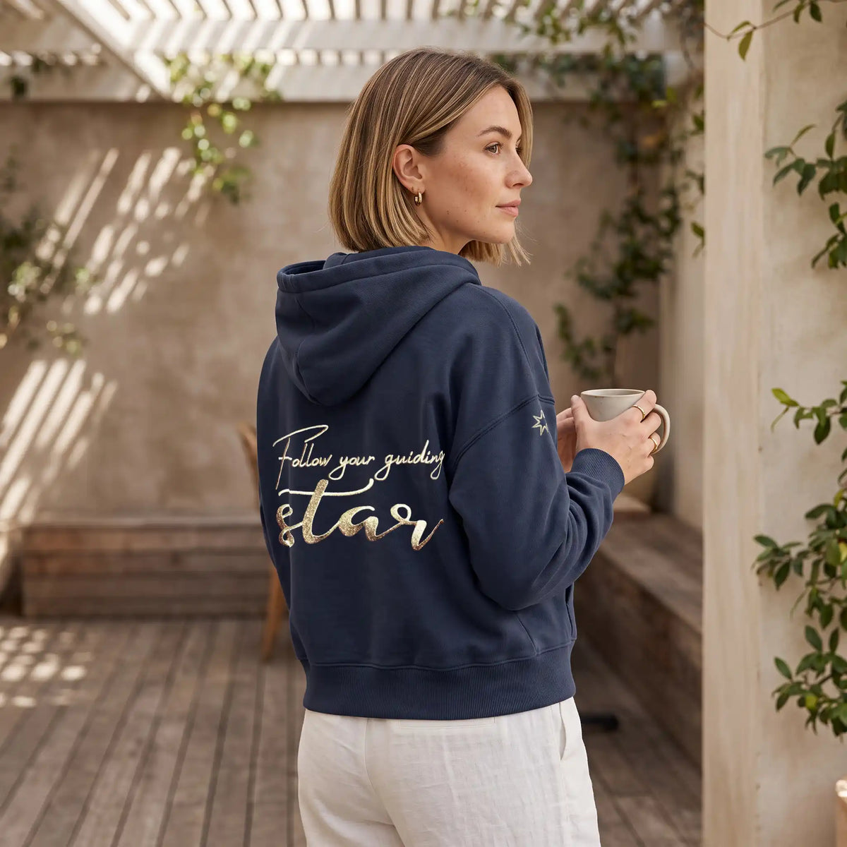 Model trägt eine dunkelblaue Damen Baumwoll Sweatjacke mit Affirmation, große Stickerei am Rücken in metallic-Gold, Affirmation 'Follow your guiding star'. Logostickerei am Ärmel in metallic-gold: siebenzackiger Stern. Sie steht auf einer schönen Terrasse.
