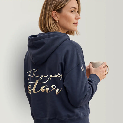 Model trägt eine Damen Hoodie Sweatjacke in dunkelblau, große Stickerei am Rücken in metallic-Gold, Affirmation 'Follow your guiding star'. Logostickerei am Ärmel in metallic-gold: siebenzackiger Stern