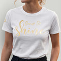 Model vor grauer Wand, sie trägt ein spirituelles Bio TShirt für Damen mit Stickerei in der Farbe weiß, die Frontstickerei schimmert golden, Schriftzug 'Time to shine'. Sternenstickerei (Logo) am rechten Ärmel.
