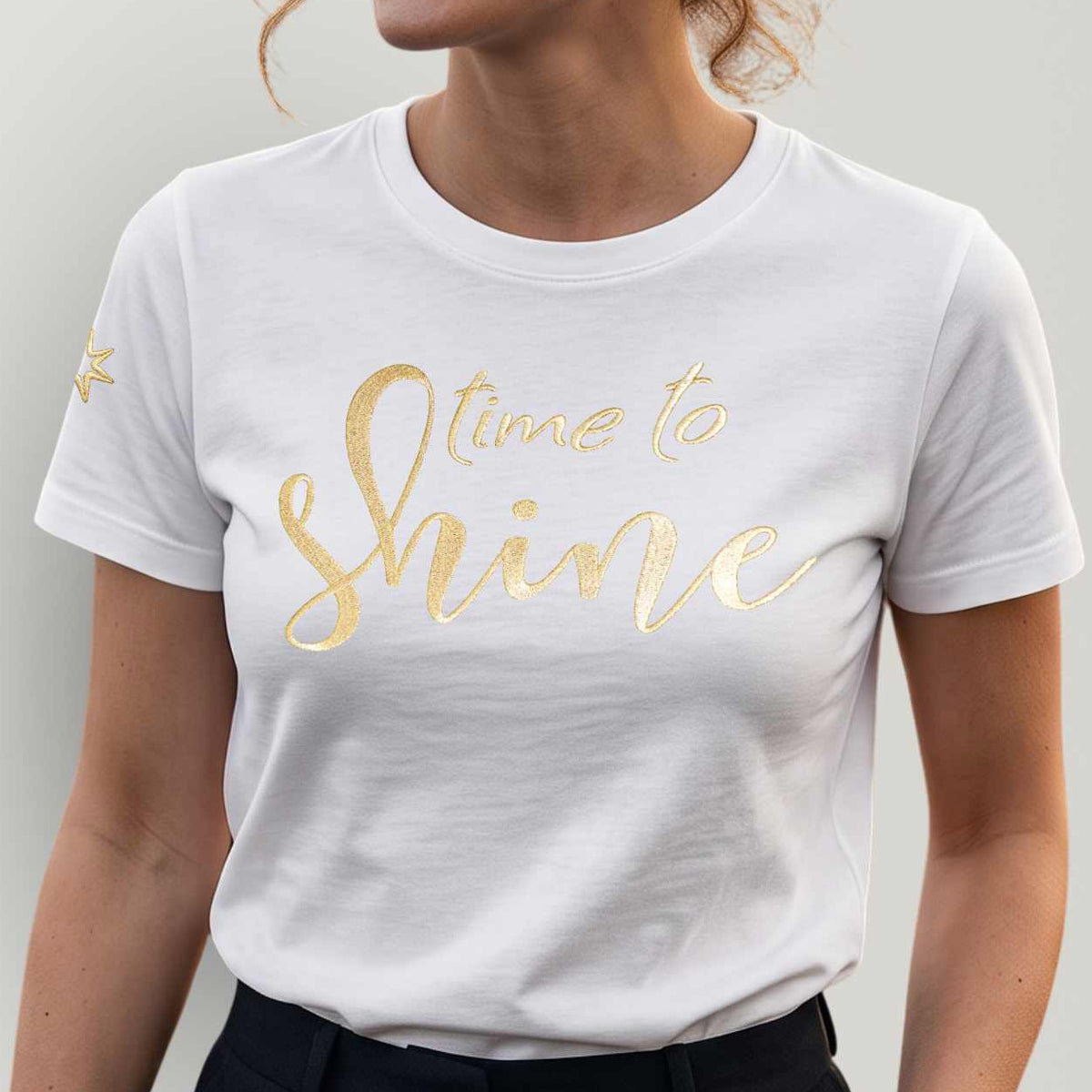 Model vor grauer Wand, sie trägt ein spirituelles Bio TShirt für Damen mit Stickerei in der Farbe weiß, die Frontstickerei schimmert golden, Schriftzug 'Time to shine'. Sternenstickerei (Logo) am rechten Ärmel.