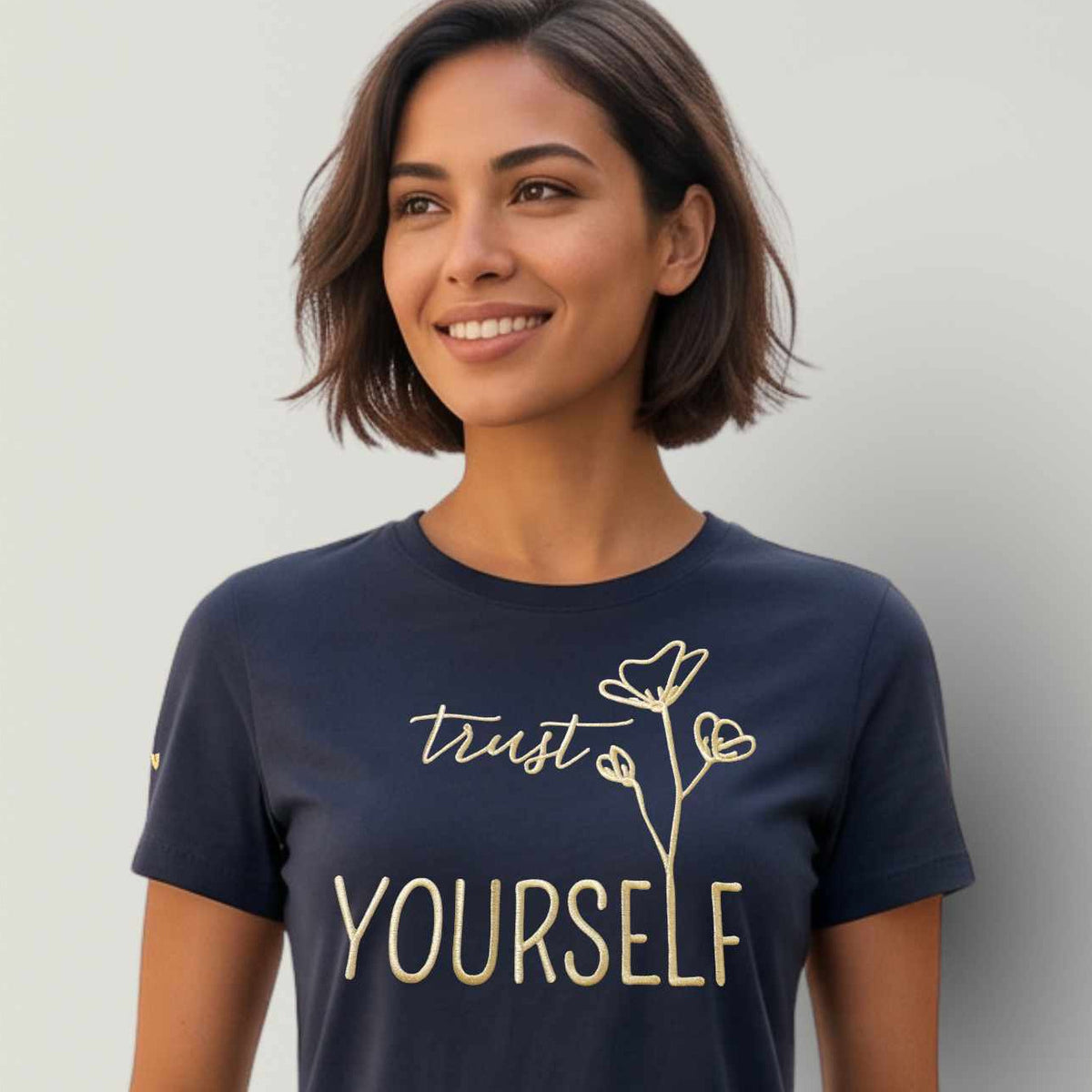 Model trägt ein dunkelblaues Damen T-Shirt aus Bio Baumwolle, mit Affirmation als große Frontstickerei in metallic-gold, Schriftzug 'Trust yourself'. Logostickerei (Stern) am rechten Ärmel. Das Model steht vor einer grauen Wand.