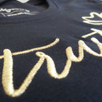 Nahaufnahme der schimmernden Goldstickerei mit dem Schriftzug 'Trust yourself' auf einem dunkelblauen Bio Damen TShirt aus reiner Baumwolle.